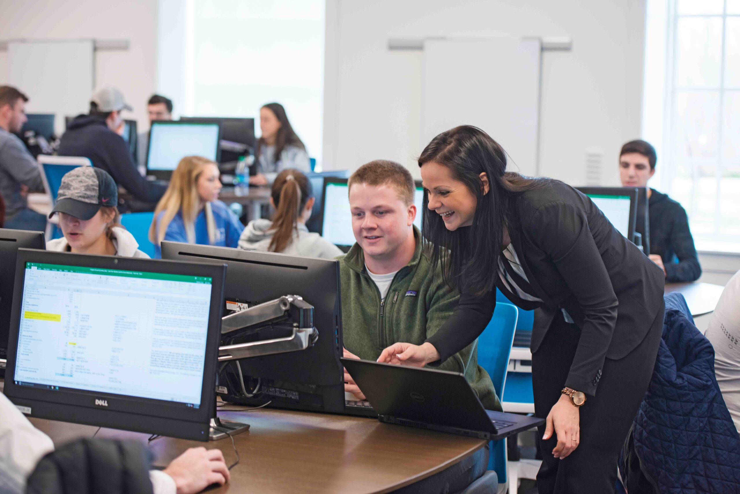 Photograph of A faculty member leans over to assist a student working at a computer in a busy classroom where other students are using desktop monitors.